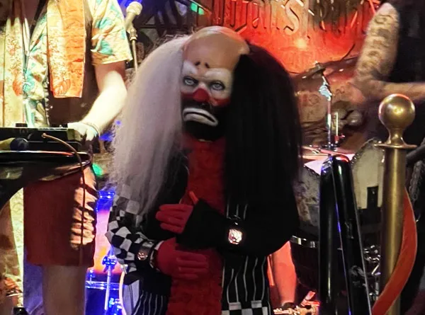 Bobbateat Fuzz in a sad-klown mask with long black and white hair, side stage