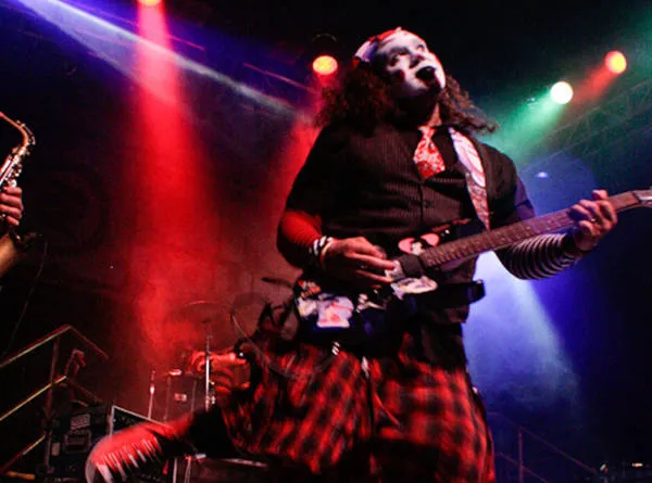 Oofoo playing guitar in a plaid kilt under red and green stage light